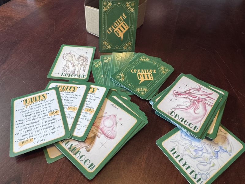 Creature Pit card game: green-backed cards and illustrated creature cards, including a unicorn, dragon, mushroom and jellyfish, spread on a wooden table with a rules card
