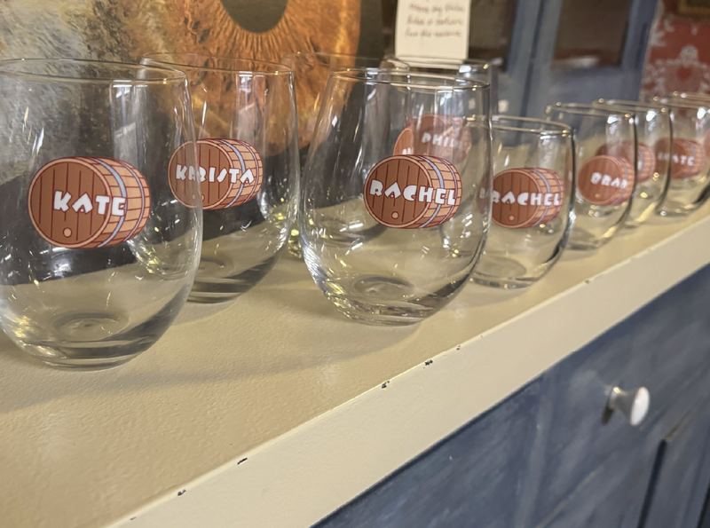 Row of stemless wine glasses on a shelf, each marked with a round barrel decal and a name