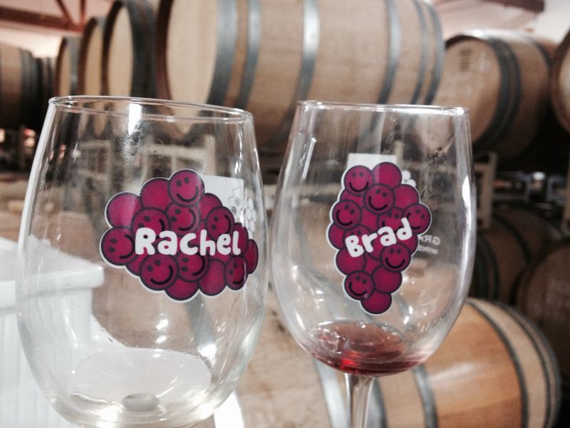 Two wine glasses in a barrel room, each with a purple smiley grape bunch decal personalized with a name