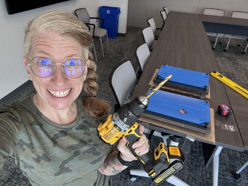 Rachel smiling and holding a power drill in a boardroom, with two blue-covered room signs lying on the table ready to install